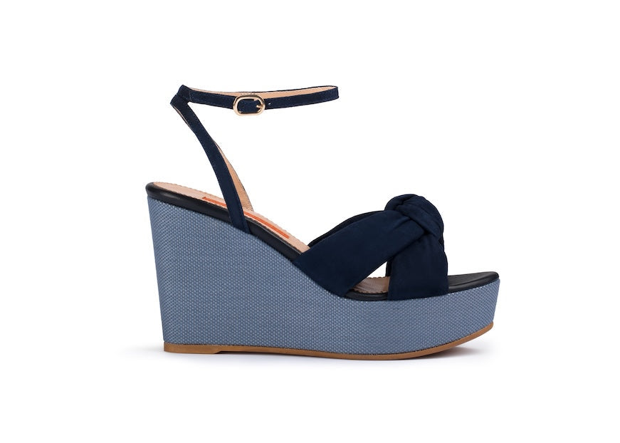 A single women's wedge sandal shown in side profile on a plain white background. The upper is dark navy blue suede with an ankle strap and a knotted detail at the toe, set on a light blue-gray platform wedge with a tan outsole.