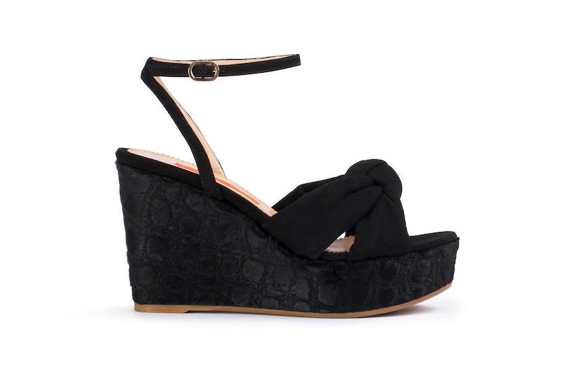 A single black wedge sandal with an ankle strap and a knotted front detail, shown in profile against a plain white background.